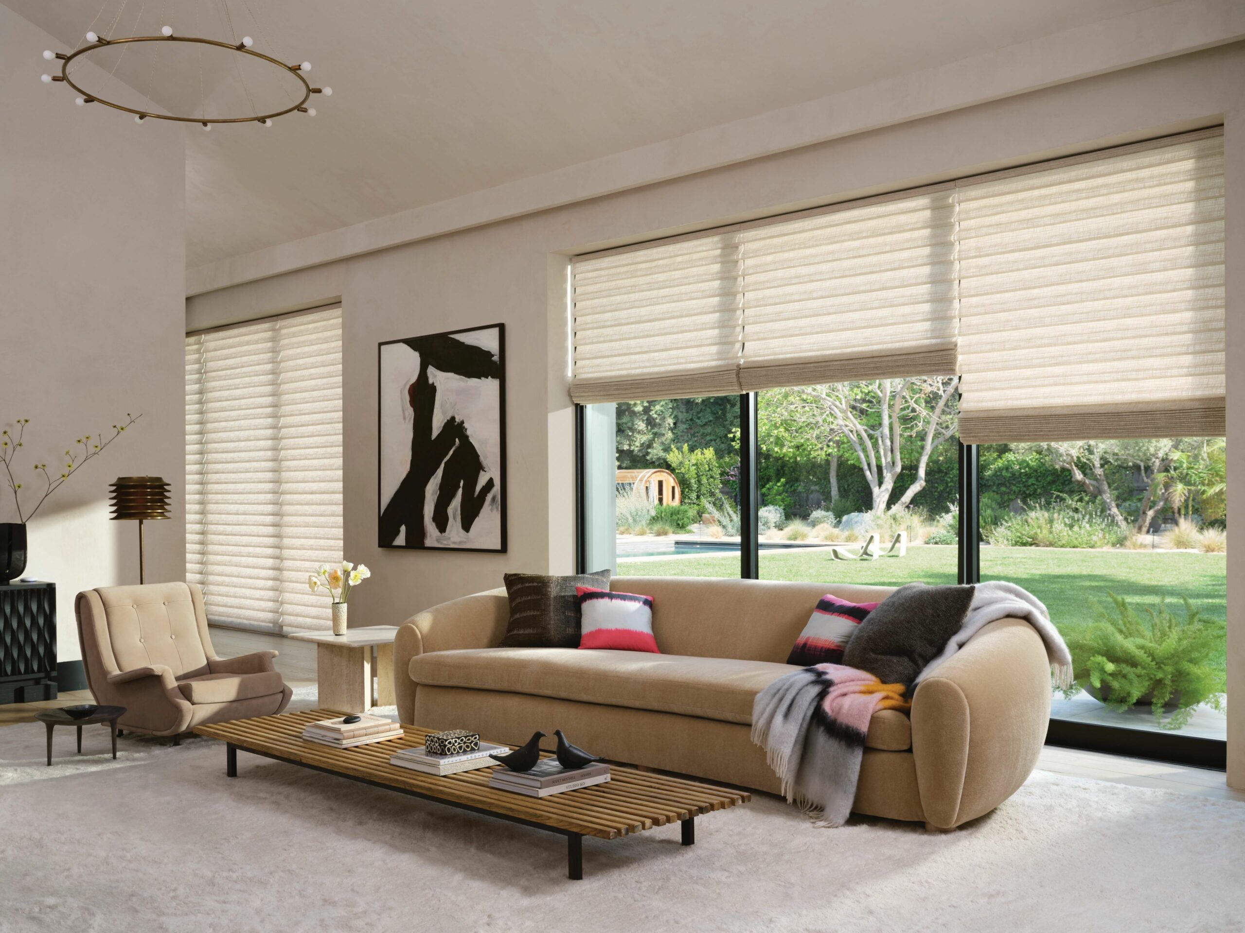 Contemporary living room with beige Roman shades covering wide windows overlooking a garden