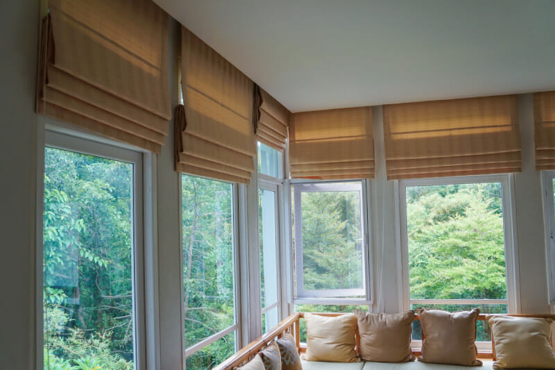Custom blinds and window treatments featuring beige Roman shades in a bright corner room with large windows and outdoor greenery views
