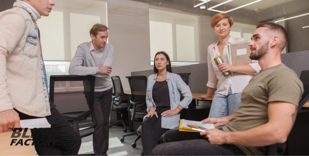 A group of office workers engaged in a discussion, standing around a desk with papers and a laptop, in a brightly lit modern office environment.